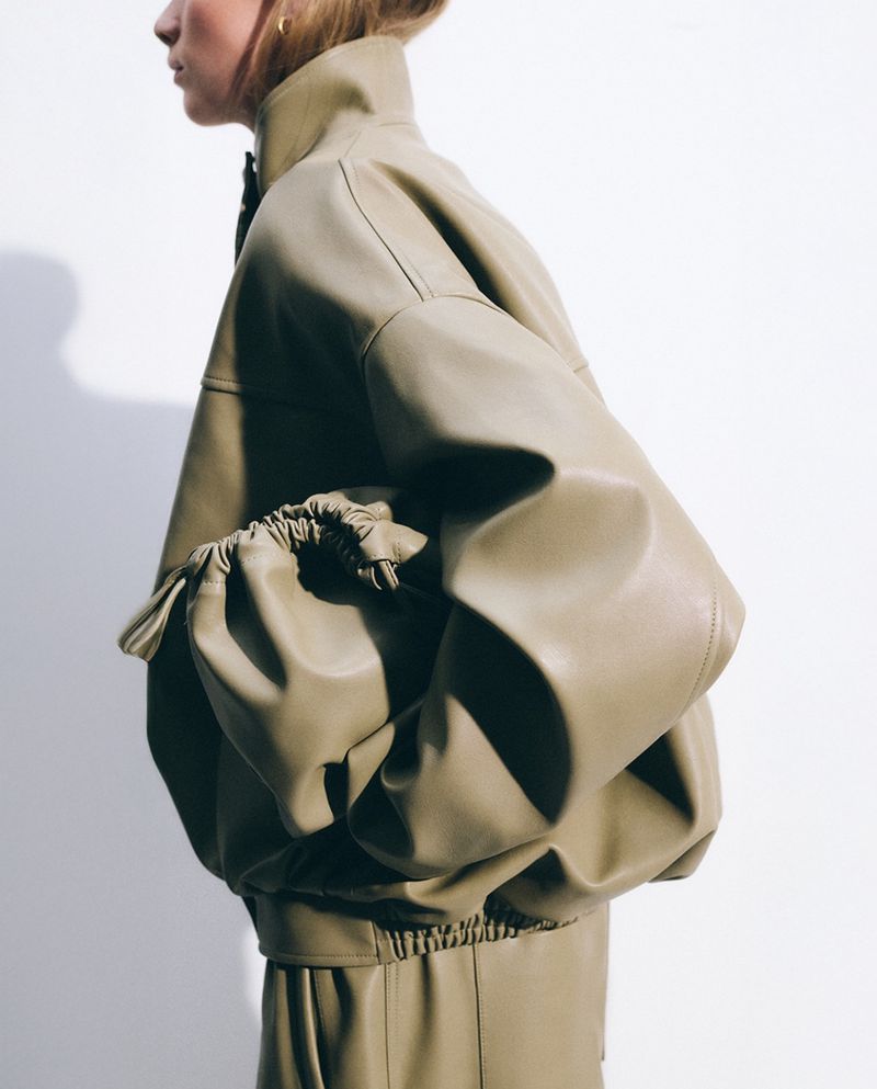 Side profile of a person wearing a beige faux leather jacket with balloon sleeves. The backdrop is a plain, neutral tone.