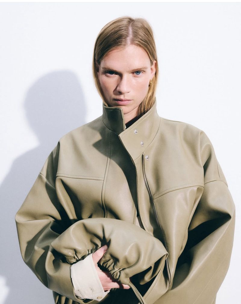 Blonde adult female poses in a studio, wearing a pale olive-green faux-leather jacket with a high collar and a neutral expression.