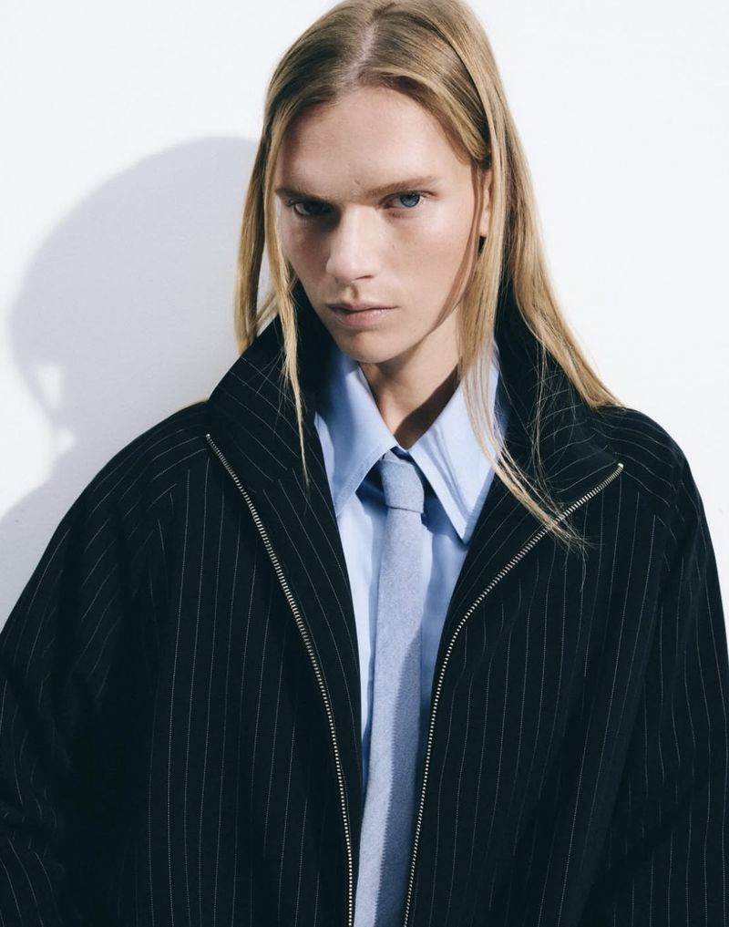 Portrait of a young adult with long blond hair wearing a pinstripe blazer, light blue shirt, and tie against a white backdrop.