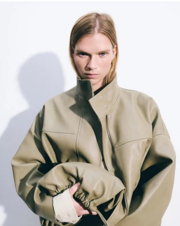 Model wearing an olive green leather jacket holding a matching pouch bag against a plain white background.