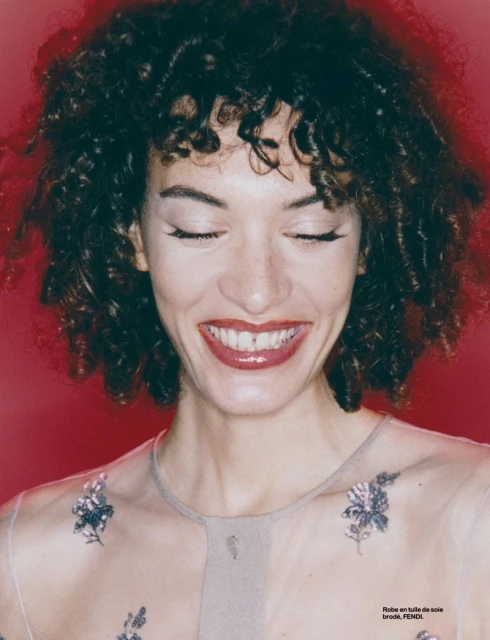 Close-up of a woman with curly dark hair, fair skin, and a bright smile wearing a sheer top with floral embroidery on a red background.