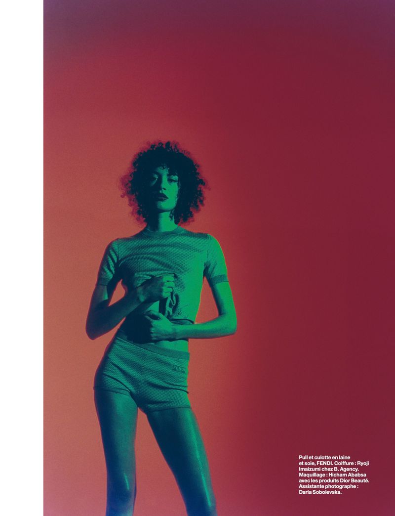 Young woman with afro poses in a green two-piece outfit against an orange backdrop with blue gel lighting in a studio fashion shot.