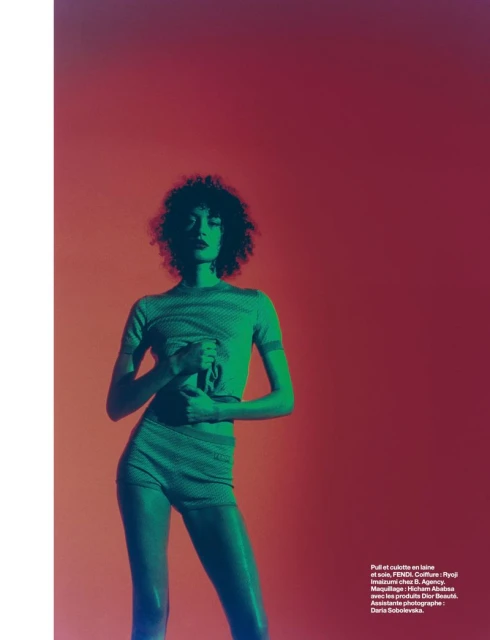 Young woman with afro poses in a green two-piece outfit against an orange backdrop with blue gel lighting in a studio fashion shot.