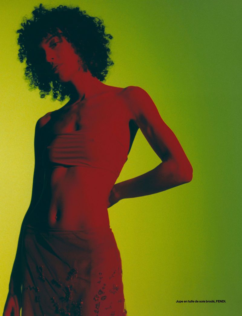 An adult female fashion model with curly hair is wearing a bandeau top and skirt, posing in front of a yellow-green studio backdrop.