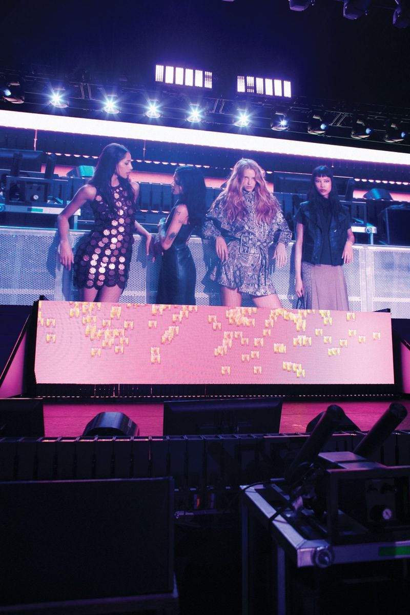 Four women are standing on a stage with an illuminated screen, wearing dresses and fashionable outfits under stage lights in an indoor venue.