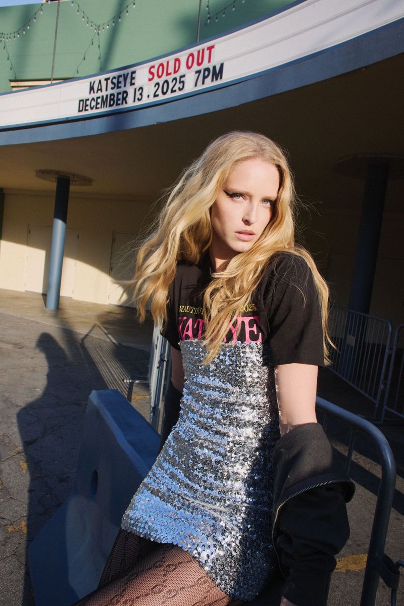 A woman with long blonde hair poses in front of a building with a marquee that reads "Katseye Sold Out December 13, 2025 7PM."