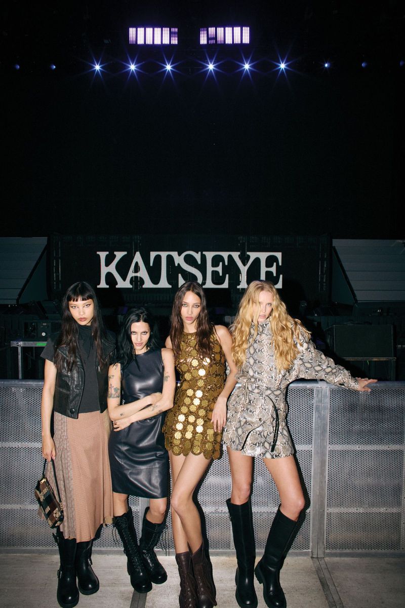 Four fashion models pose in dresses and high boots against a concert stage backdrop with stage lighting and bold typographic design.