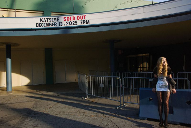 A young adult woman in a glittering dress and patterned tights poses in front of an outdoor theater with a marquee and closed metal barriers.
