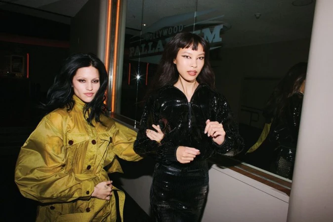 Two adult women pose in an indoor environment, wearing fashionable shiny outerwear. One wears a yellow jacket and one wears black.