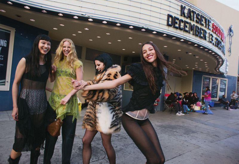 Four young women pose playfully outside a theater, dressed in sheer tops and coats. The image captures a carefree, fashionable moment.
