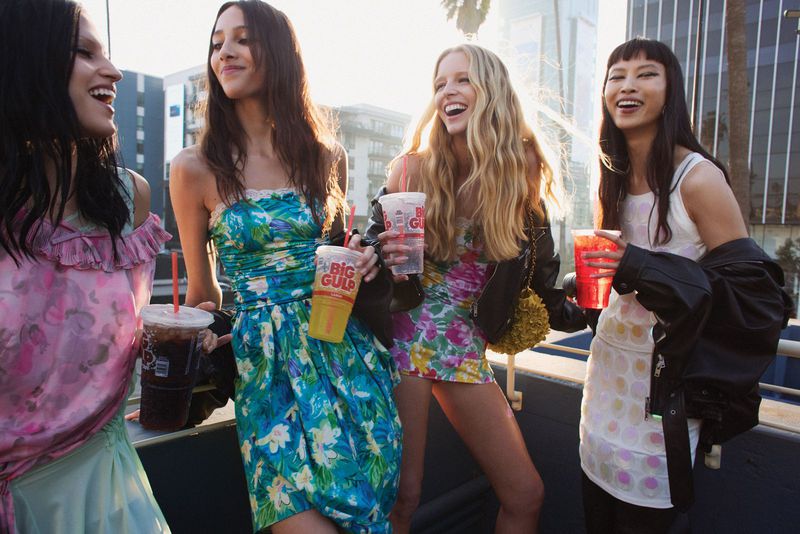 Four women in floral dresses stand outside, holding Big Gulp cups in an urban setting, smiling, with sunlight and city buildings visible.
