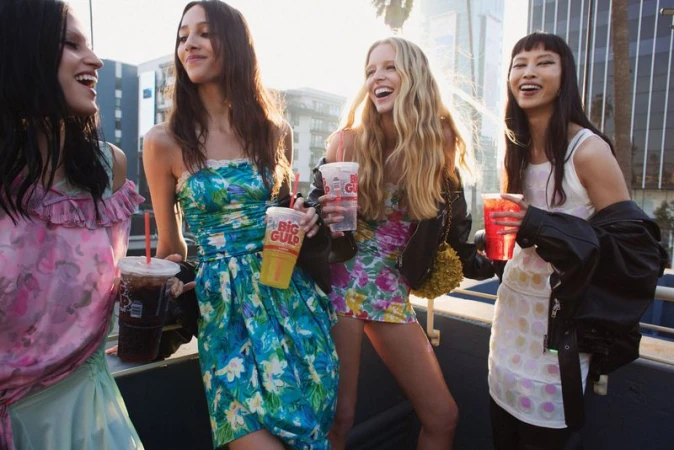 Four women in floral dresses stand outside, holding Big Gulp cups in an urban setting, smiling, with sunlight and city buildings visible.