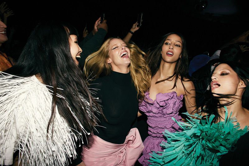 Group of stylish women laughing and posing in a dark event space, adorned with white and teal feather-like shawls and dresses.