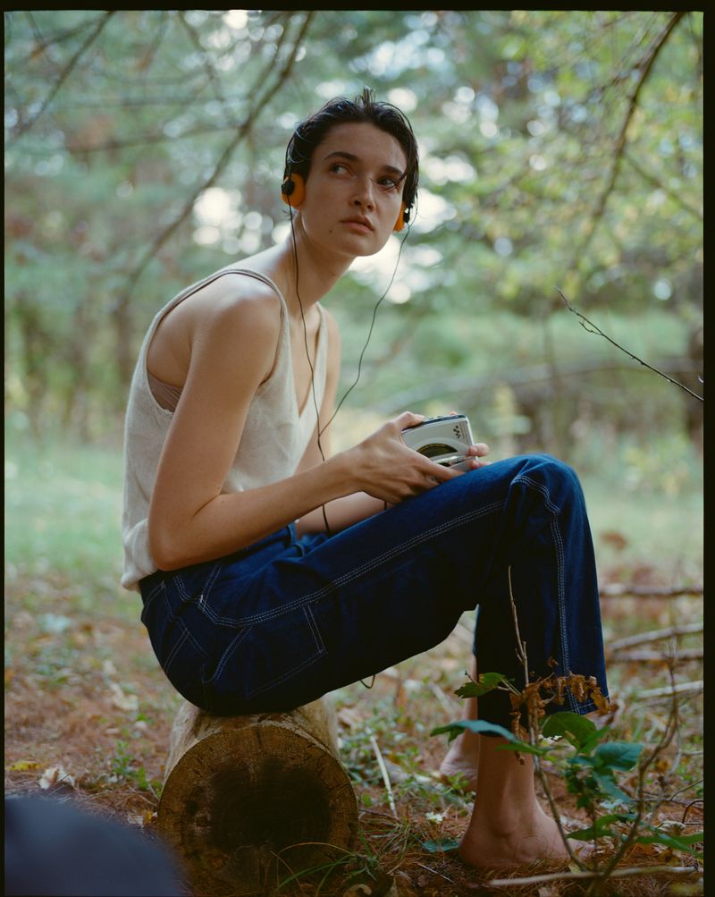 Young woman with headphones sits on a log in the forest, holding a music player. Dark blue jeans and a cream top complete the portrait.