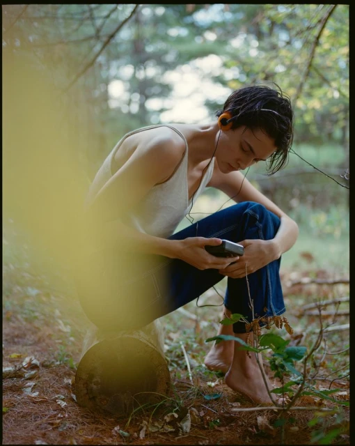 Person listening to music on a portable player in a forest, sitting on a log wearing a tank top and jeans, with blurred foreground.