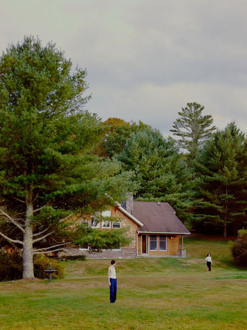 Two figures stand in a grassy field near a tan house with a brown roof, surrounded by lush green trees under a cloudy sky.