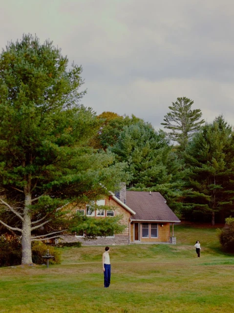 Two figures stand in a grassy field near a tan house with a brown roof, surrounded by lush green trees under a cloudy sky.
