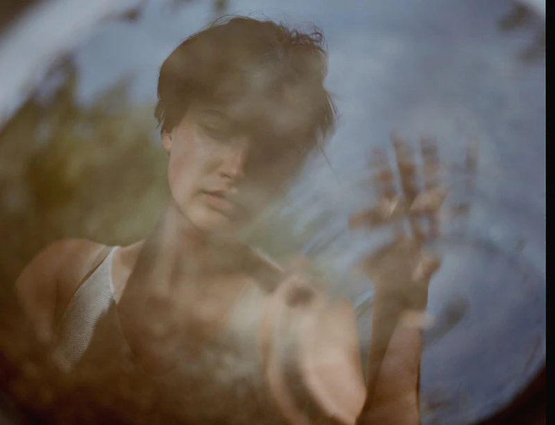 Dreamy portrait of a young woman with short brown hair and a white tank top, her hand slightly blurred and raised near her face.
