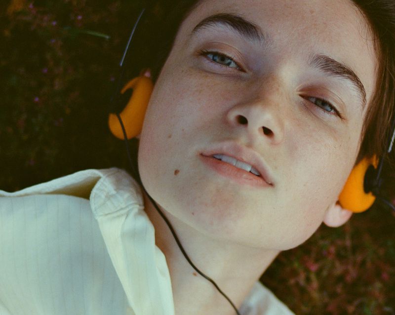 Close-up portrait of a model wearing yellow headphones and a white shirt, lying outdoors against a textured natural ground