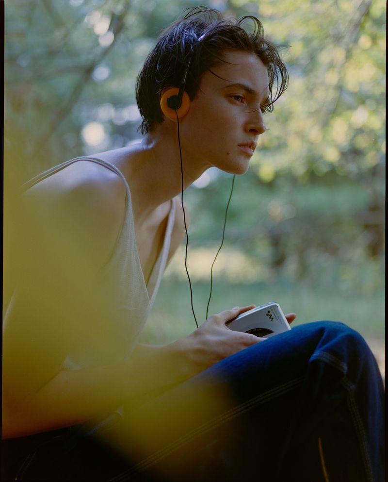 Young adult male listening to a vintage Walkman with headphones, seated outdoors in a forest environment with dappled sunlight.