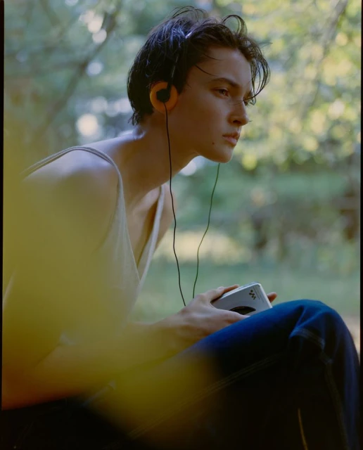 Young adult male listening to a vintage Walkman with headphones, seated outdoors in a forest environment with dappled sunlight.