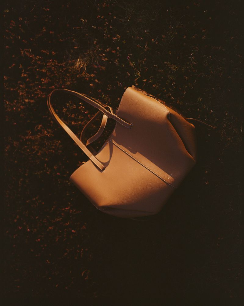 Studio shot of a stylish tan leather tote bag on a dark floral surface, bathed in warm, directional lighting.