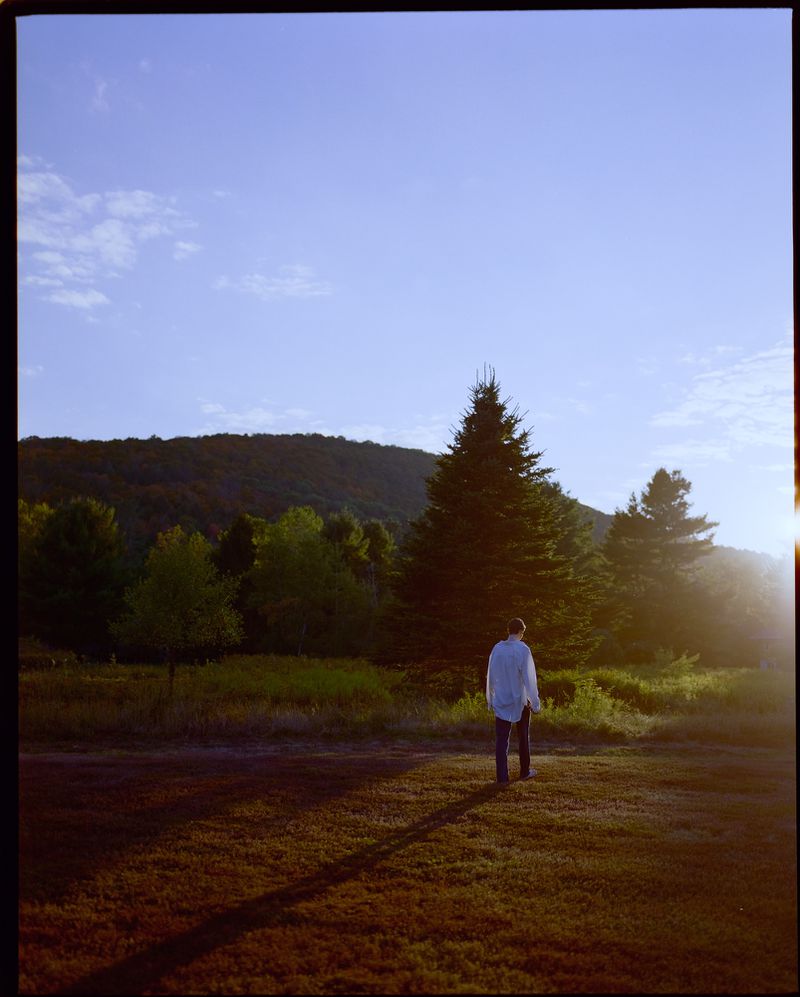 A person in a white button-down shirt walks away across a grassy meadow at sunset, casting a long shadow toward evergreen trees and mountains.