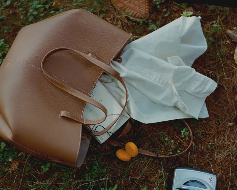 Tan leather tote bag with a white shirt, book, headphones, and Walkman sitting on a grassy surface in an outdoor setting. 