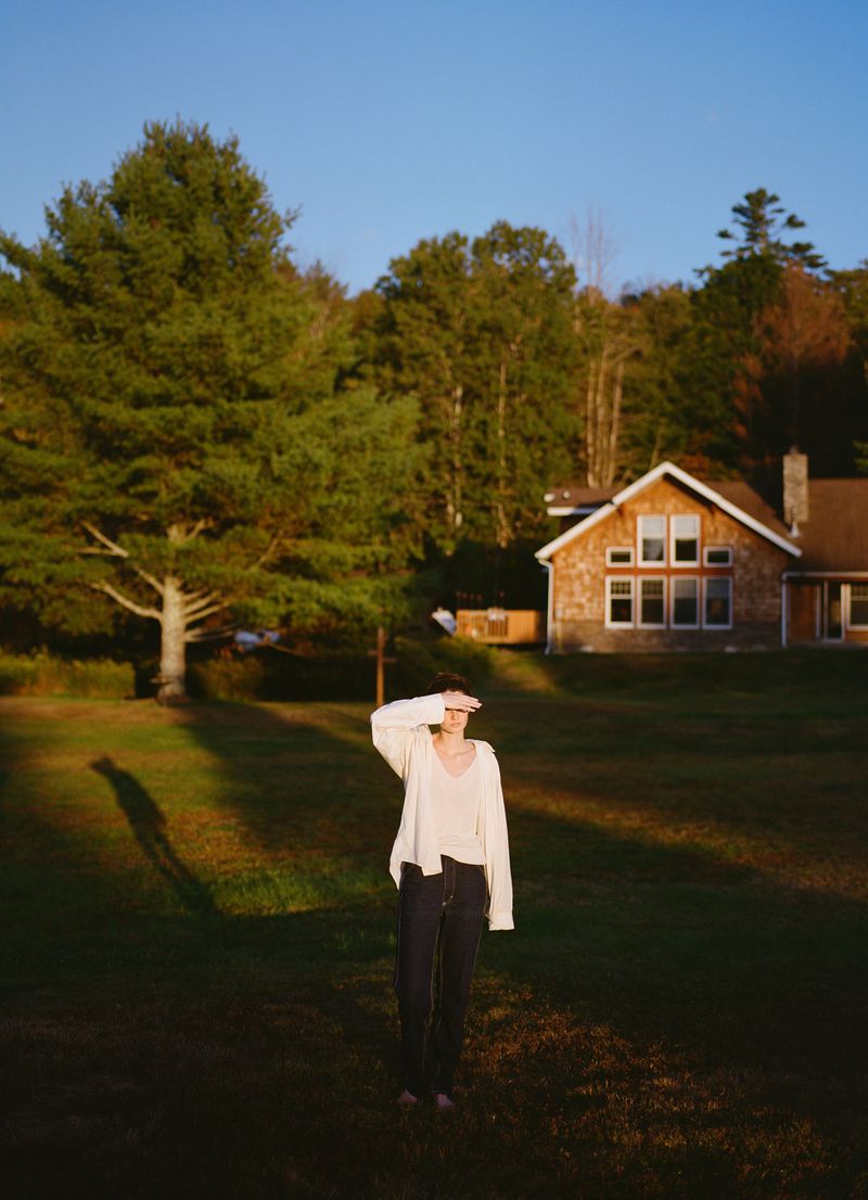 A person stands in a grassy field, shielding their eyes from the sun, with a quaint house and lush green trees in the background.