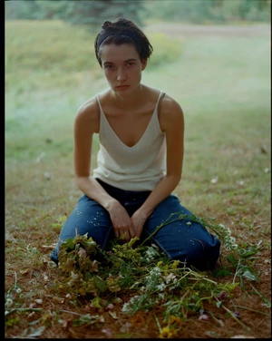 A woman kneeling on grass in a white tank top and blue denim jeans amidst scattered wildflowers and foliage.
