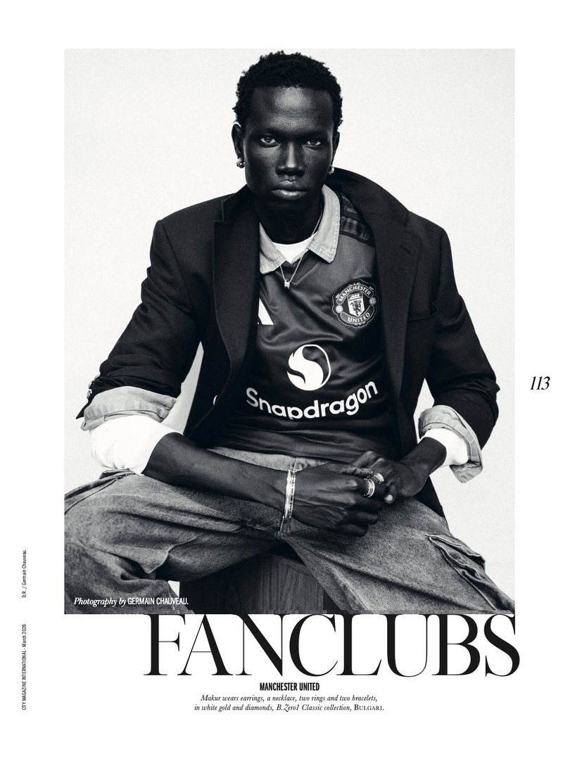 Monochromatic portrait of a Black man wearing a Manchester United jersey and blazer, seated against a bright background in a studio.