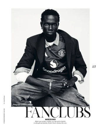 Monochromatic portrait of a Black man wearing a Manchester United jersey and blazer, seated against a bright background in a studio.