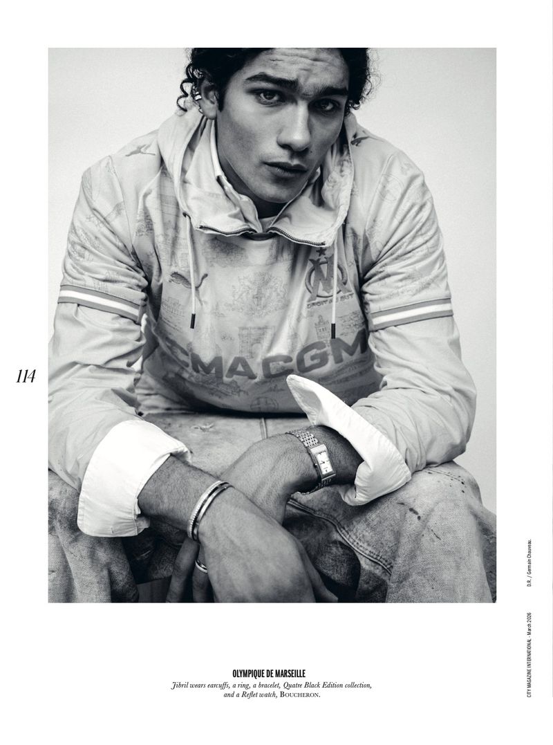 Black-and-white portrait of a dark-haired male wearing a branded hooded top and bracelets, gazing intently at the viewer, studio lit.