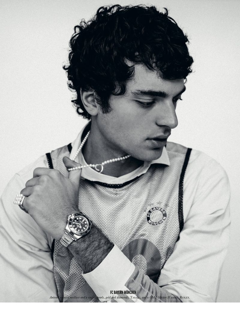 Black and white portrait of a stylish young adult male model with dark curly hair wearing jewelry and a collared shirt indoors.