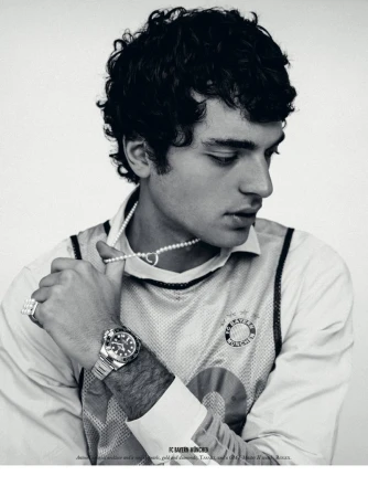 Black and white portrait of a stylish young adult male model with dark curly hair wearing jewelry and a collared shirt indoors.
