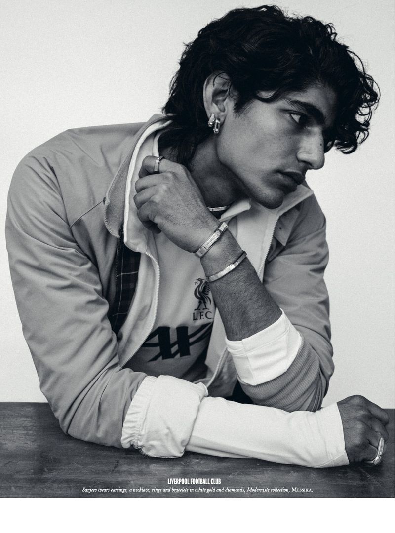 Portrait of a young adult man in a Liverpool Football Club jersey and jacket, wearing jewelry, set against a neutral background.