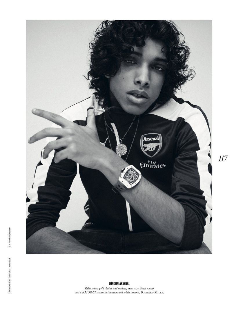 Monochrome portrait of a young adult man with curly hair wearing a track jacket, watch, and necklaces against a light backdrop.