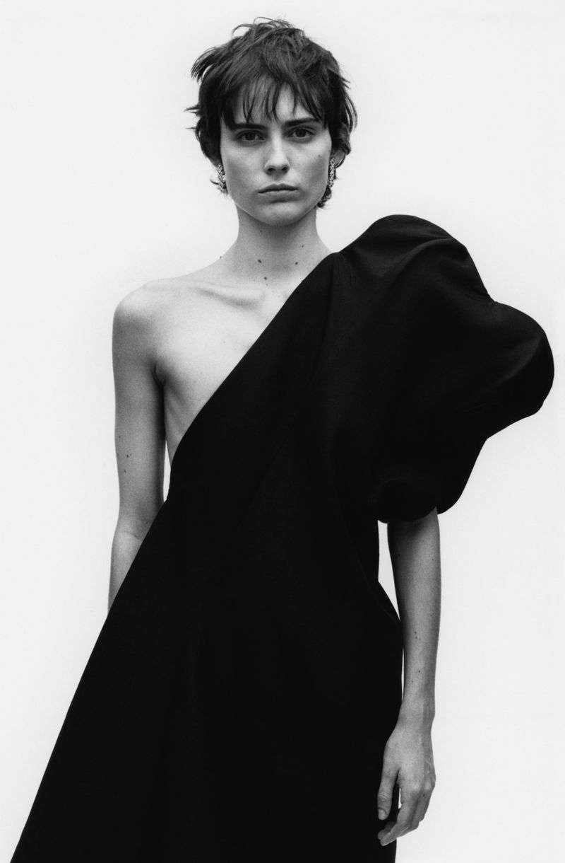 Black and white portrait of a woman with short dark hair wearing a dramatic black dress against a plain white background.