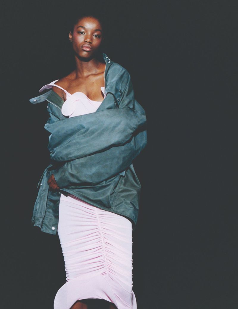Elegant Black woman in a pink dress and oversized jacket against a dark backdrop, showcasing fashion and style.