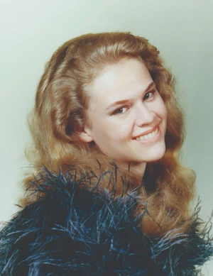 Portrait of a smiling woman with wavy blonde hair wearing a dark feather-trimmed garment against a pale green backdrop.