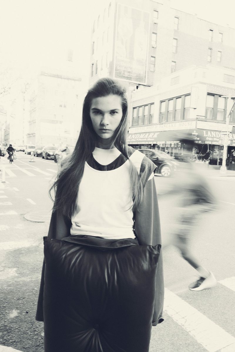 Teen girl in a stylish, oversized black outfit poses on a city street with cars and pedestrians visible in the monochromatic background.