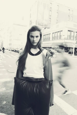 Teen girl in a stylish, oversized black outfit poses on a city street with cars and pedestrians visible in the monochromatic background.