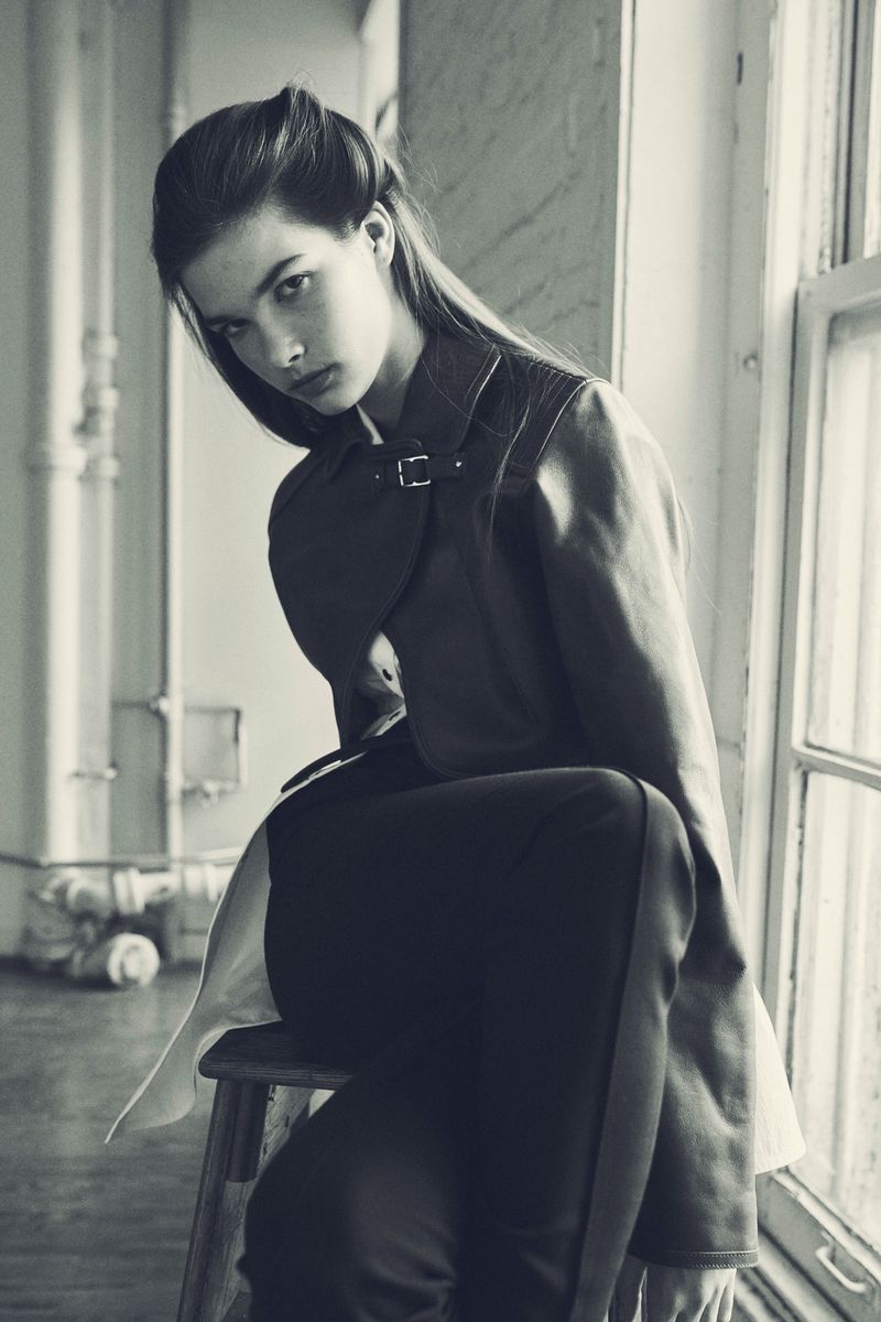 Monochrome portrait of a young woman in a jacket sitting on a wooden stool near a window with natural light, showcasing fashion style.