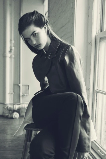 Monochrome portrait of a young woman in a jacket sitting on a wooden stool near a window with natural light, showcasing fashion style.