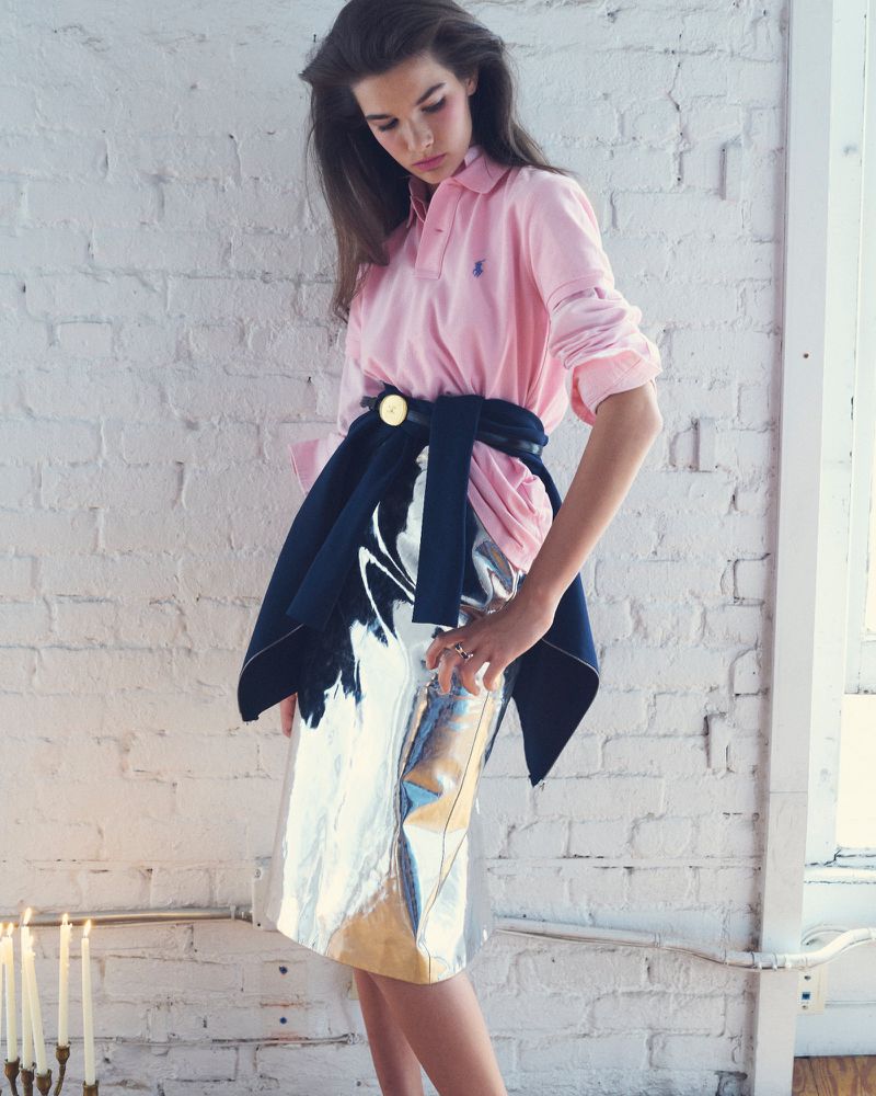 An adult woman poses indoors wearing a light pink polo shirt, a navy blue jacket tied around her waist, and a shiny silver skirt.