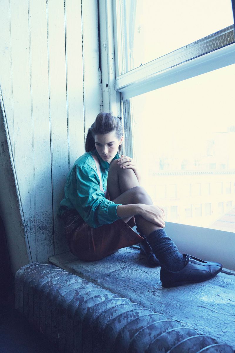 Young woman in a teal shirt sits by a window with dark socks and shoes, providing a soft fashion editorial image in indoor lighting.