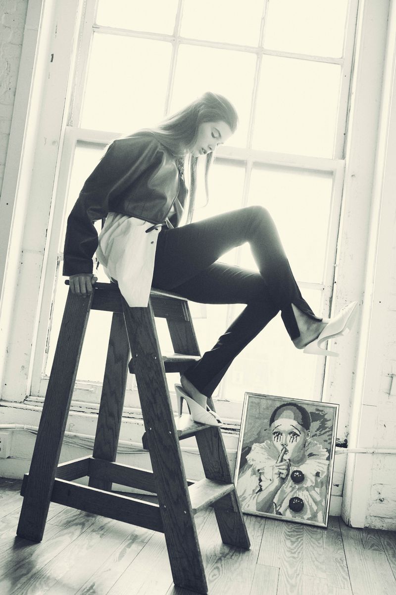 A fashionable young woman sits on a wooden ladder in front of a bright window, posing next to a framed clown painting, in a photo studio.