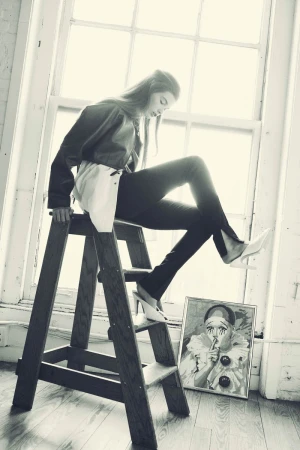 A fashionable young woman sits on a wooden ladder in front of a bright window, posing next to a framed clown painting, in a photo studio.