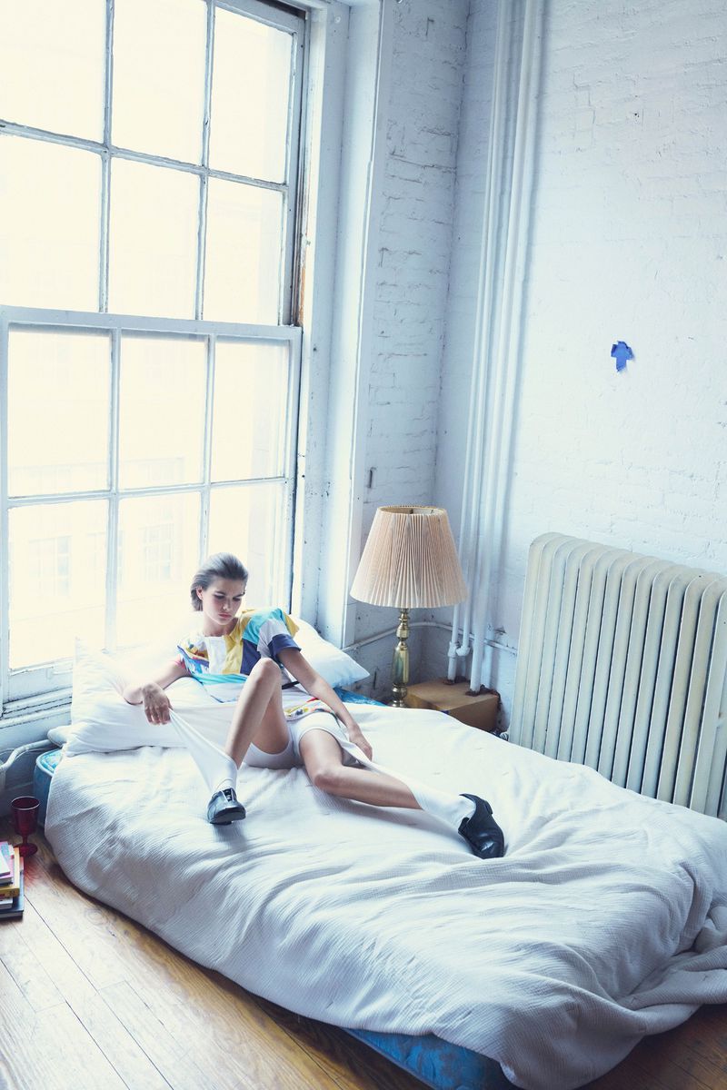 Young woman in a colorful top and white shorts relaxes on a white bed in a sunlit room near a large window, creating a casual, relaxed scene.
