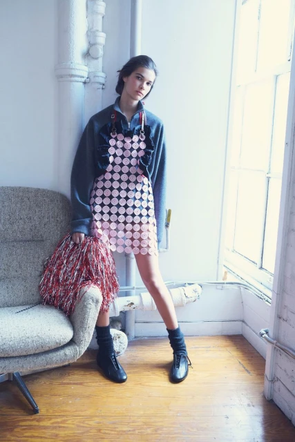 Young woman in fashionable pink circle dress and gray sweatshirt, posing with a red and silver pom-pom, near a chair and window.